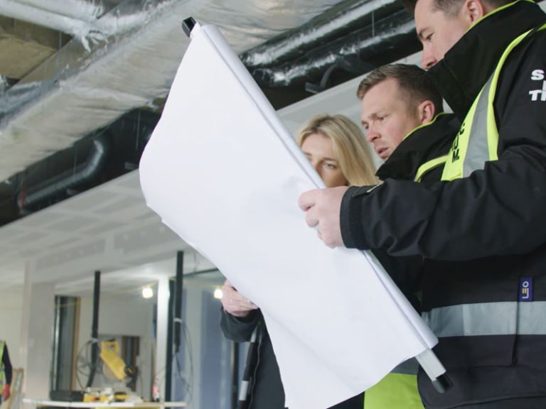Three people look at plans for a workplace whilst on site